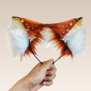 May include: A headband with fox ear design. The ears are made of orange, brown, and white faux fur, decorated with gold details and small pearl-like adornments. The headband is held by a hand against a plain backdrop.