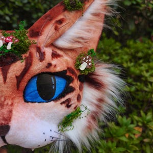 May include: A close-up of a cat-like mask with a brown and orange spotted pattern, blue eyes, and white whiskers. The mask is decorated with green moss and small mushroom accents. The ear has white and brown fur.