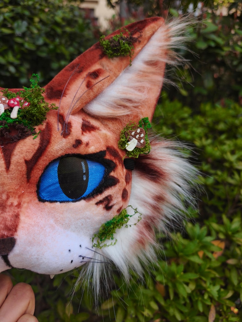 May include: A close-up of a cat-like mask with a brown and orange spotted pattern, blue eyes, and white whiskers. The mask is decorated with green moss and small mushroom accents. The ear has white and brown fur.