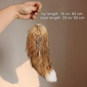 May include: A brown and white faux fur tail keychain with a gold-coloured clasp and decorative charms. The fur length is 40 cm, and the total length is 50 cm.