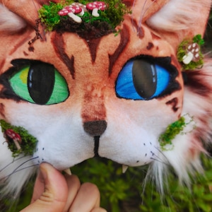 May include: A close-up of a cat mask with one green eye and one blue eye. The mask is brown with darker brown stripes and is decorated with green moss and small red and white mushroom accents. The mask has white whiskers and small fangs.