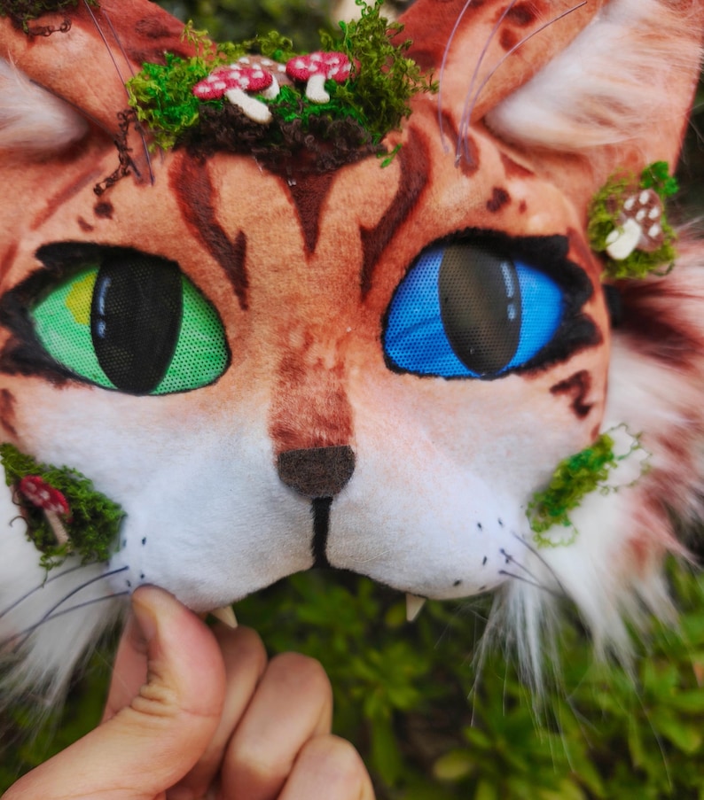 May include: A close-up of a cat mask with one green eye and one blue eye. The mask is brown with darker brown stripes and is decorated with green moss and small red and white mushroom accents. The mask has white whiskers and small fangs.