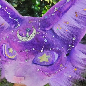 May include: A purple plush cat mask featuring long ears and faux fur accents. The surface is decorated with silver star and crescent moon charms, clear rhinestones, and small glitter particles. The eyes are mesh with yellow star designs.