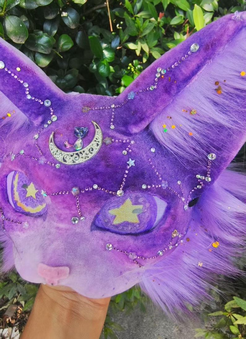 May include: A purple plush cat mask featuring long ears and faux fur accents. The surface is decorated with silver star and crescent moon charms, clear rhinestones, and small glitter particles. The eyes are mesh with yellow star designs.