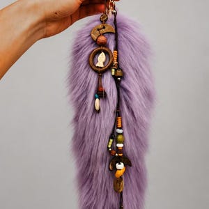 May include: A lavender faux fur tail keychain featuring a gold-toned metal clasp. The accessory is decorated with two dark wooden crescent moon charms, a small wooden ring containing a white fish charm, and various colorful beads.