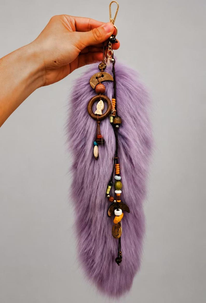 May include: A lavender faux fur tail keychain featuring a gold-toned metal clasp. The accessory is decorated with two dark wooden crescent moon charms, a small wooden ring containing a white fish charm, and various colorful beads.