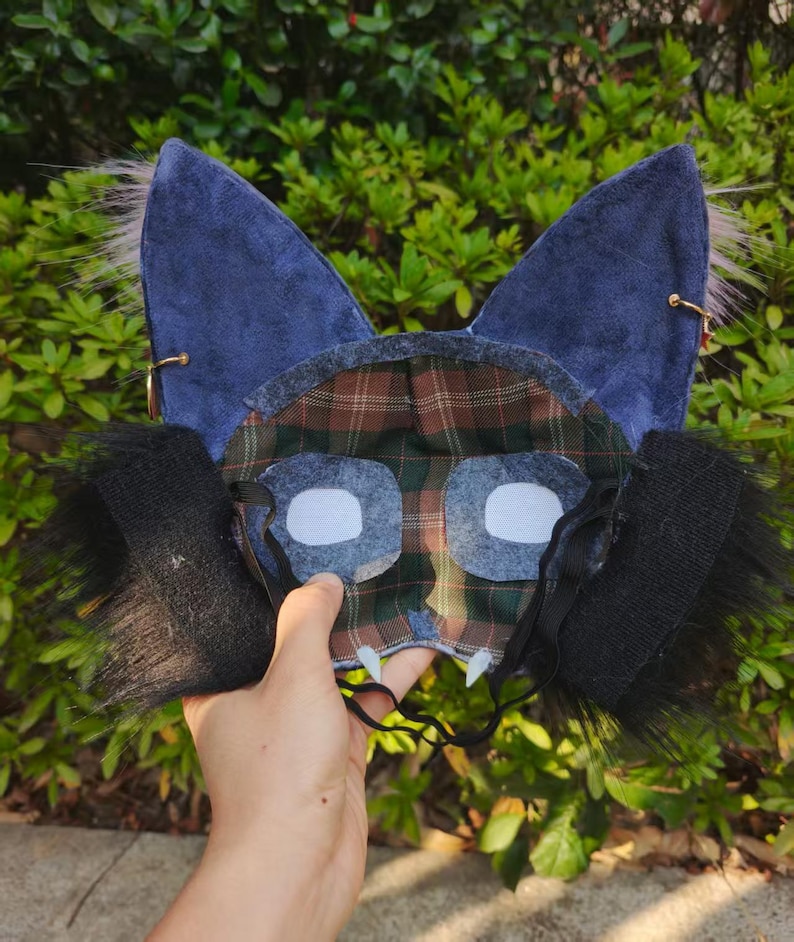 May include: A blue and plaid fabric mask with pointed ears, white eye cutouts, and black fur accents. The mask has a plaid pattern on the inside and is held by a hand against a green leafy background.
