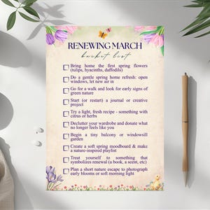 Puede incluir: Una lista impresa titulada "Renewing March Bucket List" con ilustraciones florales y de mariposas. La lista incluye actividades temáticas de primavera como traer flores a casa, refrescar el hogar y comenzar un proyecto creativo. Un bolígrafo de madera descansa a la derecha.