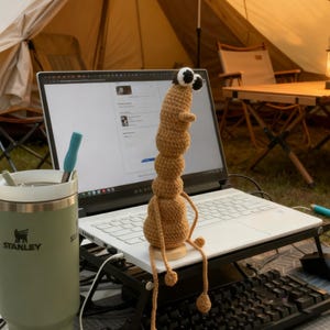 May include: A tan crocheted worm-like creature with large googly eyes sits on a laptop. A green Stanley tumbler with a silver lid and a blue straw is to the left. A tent and camping table are in the background, suggesting an outdoor workspace.