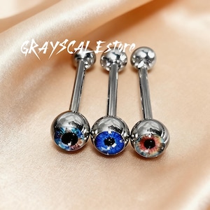 May include: Three silver-colored tongue piercings with a ball on each end. The balls feature a realistic eye design with blue, red, and black irises. The text "GRAYSCALEstore" is above the piercings.