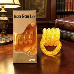 Op de afbeelding: Een doorzichtig, geel puzzel in de vorm van een gebalde vuist. De puzzel staat naast de kartonnen verpakking, waarop "Rao Rao Le" en "Manufactured in Shenzhen, China" staat. De verpakking geeft ook aan dat het product geschikt is voor personen van 15 jaar en ouder.