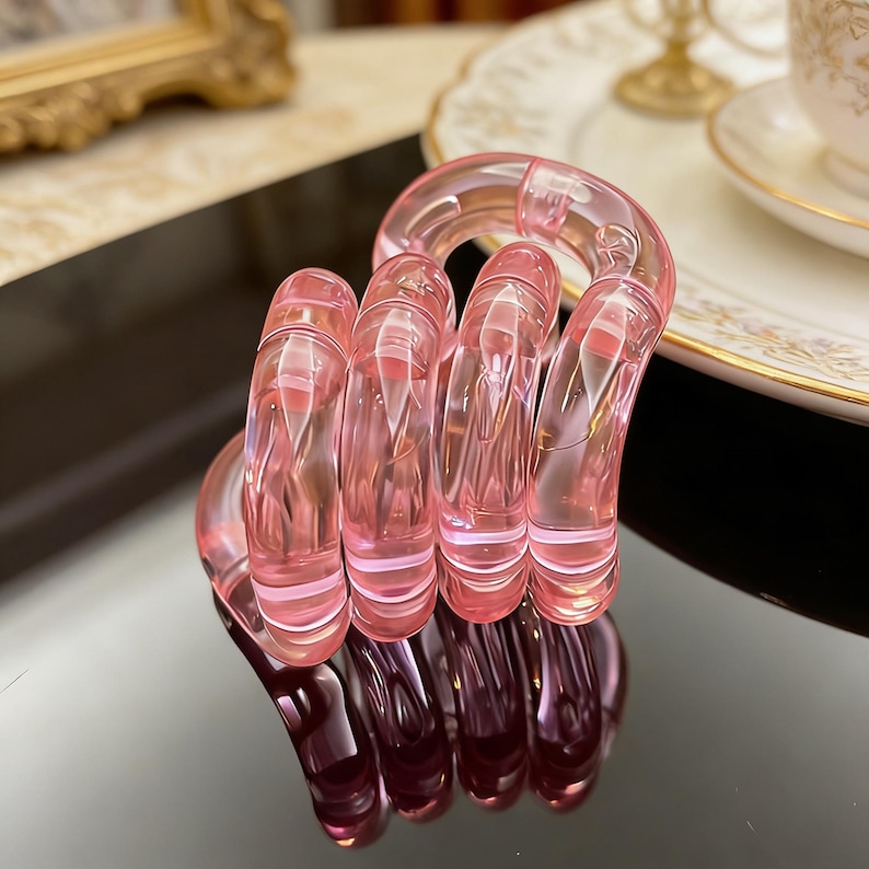 Op de afbeelding: Een roze, transparante, spiraalvormige haarband. De haarband is gemaakt van een helder, glanzend materiaal en reflecteert licht. Het ontwerp heeft een reeks verbonden, gebogen segmenten. De achtergrond bevat een decoratieve plaat en een goudomrand object.