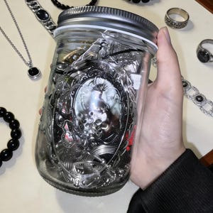 Puede incluir: Un tarro de cristal transparente lleno de varias joyas plateadas, incluyendo collares, pulseras y anillos. El tarro tiene una tapa plateada y lo sostiene una persona. Otras piezas de joyería están dispersas alrededor del tarro.