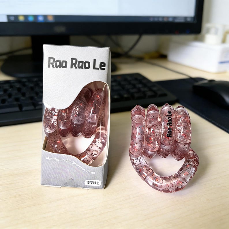 Op de afbeelding: Een doorzichtig, roze handvormig speelgoed met rode glitter, naast de doos. Op de doos staat "Rao Rao Le" en "Vervaardigd in Shenzhen, China". Het speelgoed is ontworpen om vast te houden en te manipuleren.
