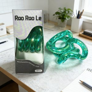 Op de afbeelding: Een groene, doorschijnende Rao Rao Le tangle-speelgoed, met een doos met de tekst "Rao Rao Le" in zwarte letters. De doos vermeldt ook "Vervaardigd in Shenzhen, China" en "15 jaar en ouder". Het tangle-speelgoed is gemaakt van verbonden, gebogen stukken.
