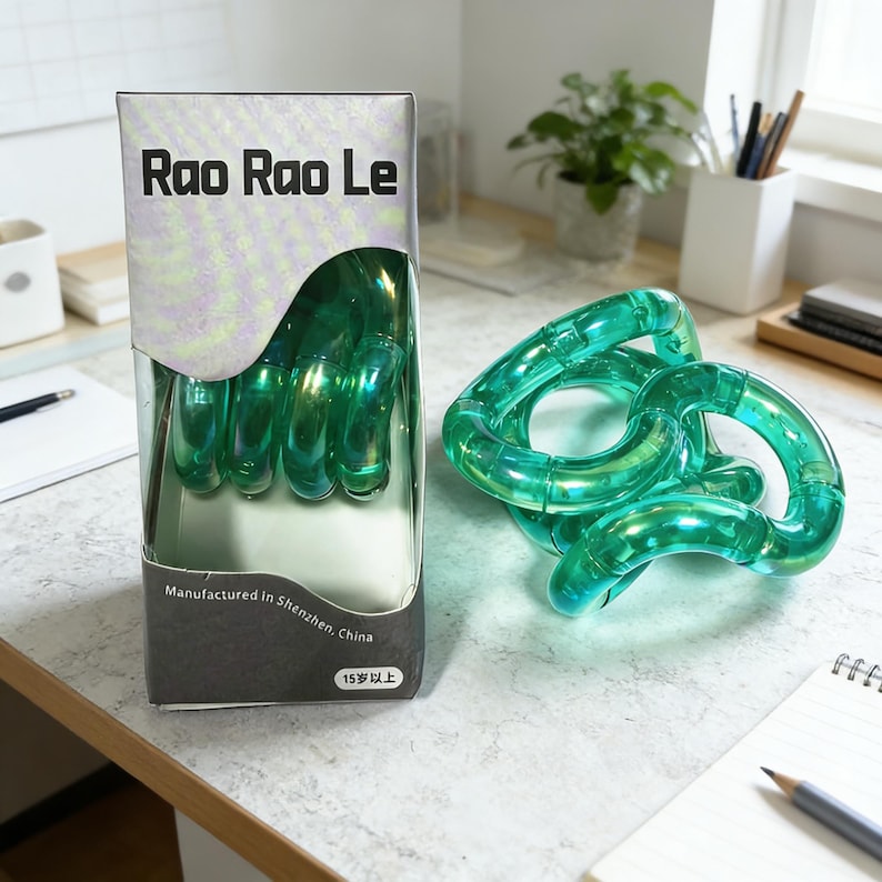 Op de afbeelding: Een groene, doorschijnende Rao Rao Le tangle-speelgoed, met een doos met de tekst "Rao Rao Le" in zwarte letters. De doos vermeldt ook "Vervaardigd in Shenzhen, China" en "15 jaar en ouder". Het tangle-speelgoed is gemaakt van verbonden, gebogen stukken.