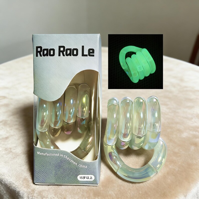 Op de afbeelding: Een doorzichtig, iriserend handvormig speelgoed met een doos met de tekst "Rao Rao Le" en "Vervaardigd in Shenzhen, China". Een kleinere afbeelding toont het speelgoed groen oplichtend. De doos heeft ook de tekst "15 jaar en ouder".