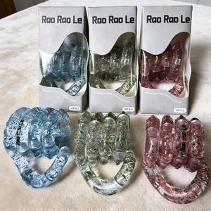Op de afbeelding: Drie vuistvormige stressspeeltjes van helder plastic, elk met glitter en een andere tint: blauw, helder en rood. De speeltjes zitten in dozen met de tekst "Rao Rao Le" en "15+ jaar".