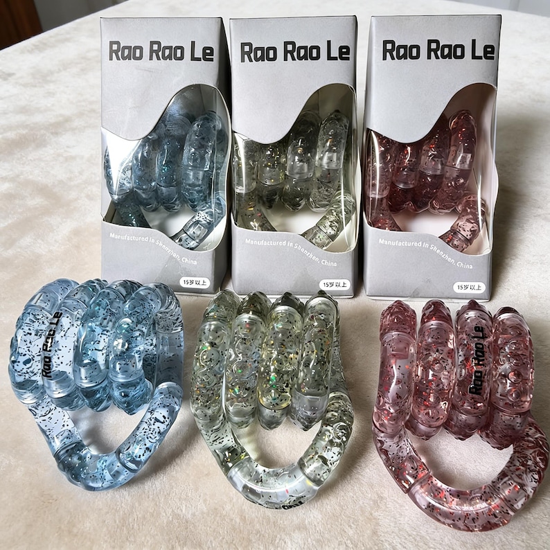 Op de afbeelding: Drie vuistvormige stressspeeltjes van helder plastic, elk met glitter en een andere tint: blauw, helder en rood. De speeltjes zitten in dozen met de tekst "Rao Rao Le" en "15+ jaar".