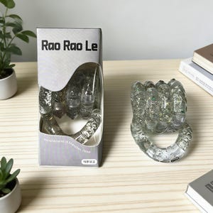 Op de afbeelding: Doorzichtig plastic handvormig fidget-speelgoed met glitter erin, naast de verpakking. Op de verpakking staat "Rao Rao Le" en "Manufactured in Shenzhen, China". Het speelgoed is ontworpen voor kinderen van 15 jaar en ouder.