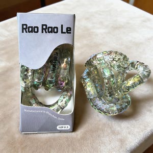 Op de afbeelding: Een doorzichtig, iriserend Tangle-speelgoed en de verpakking. Op de verpakking staat "Rao Rao Le" in grote zwarte letters. Het speelgoed is gemaakt van verbonden, gebogen stukken. De verpakking vermeldt ook "Vervaardigd in Shenzhen, China" en "15+".