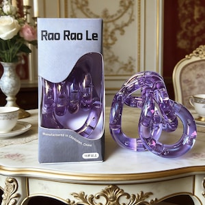 Op de afbeelding: Een paars Rao Rao Le puzzelspeelgoed, met een doos met de tekst "Rao Rao Le" en "Vervaardigd in Shenzhen, China". De puzzel is een reeks in elkaar grijpende ringen. De doos toont de puzzel in de vorm van een hand.