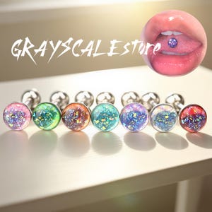 May include: A collection of colorful, sparkly tongue piercings with silver posts. The image includes a close-up of a mouth with a tongue piercing and the text "GRAYSCALESTORE". The piercings feature various colors, including pink, green, and blue.