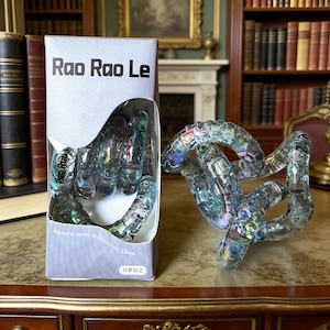 Op de afbeelding: Een doorzichtig, iriserend Rao Rao Le fidget-speelgoed. Het speelgoed bestaat uit verbonden, gebogen segmenten. De doos heeft de tekst "Rao Rao Le" en "Manufactured in Shenzhen, China". Het speelgoed staat op een houten oppervlak.