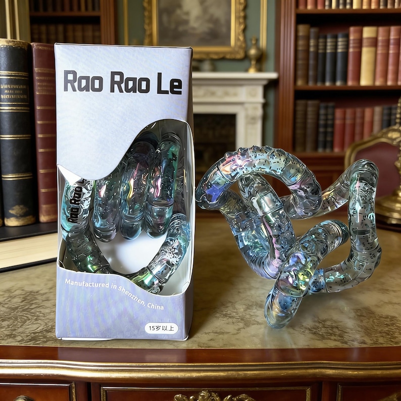 Op de afbeelding: Een doorzichtig, iriserend Rao Rao Le fidget-speelgoed. Het speelgoed bestaat uit verbonden, gebogen segmenten. De doos heeft de tekst "Rao Rao Le" en "Manufactured in Shenzhen, China". Het speelgoed staat op een houten oppervlak.