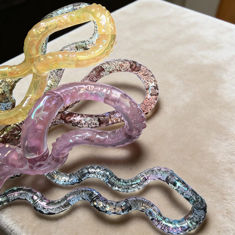 Op de afbeelding: Close-up van verschillende kleurrijke, onderling verbonden, plastic fidget-speeltjes. De speeltjes zijn in tinten geel, roze en zilver, met een glitterachtig, iriserend effect. De speeltjes zijn gerangschikt op een zacht, beige oppervlak.