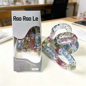Op de afbeelding: Een doorzichtig plastic fidget-speelgoed met glitter en kleurrijke segmenten wordt getoond naast de verpakking. De verpakking leest "Rao Rao Le" en "Vervaardigd in Shenzhen, China". Het speelgoed is een wirwar van verbonden, gebogen stukken.