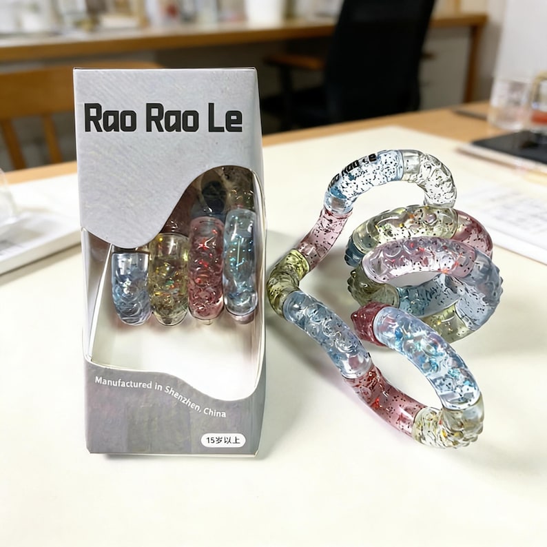 Op de afbeelding: Een doorzichtig plastic fidget-speelgoed met glitter en kleurrijke segmenten wordt getoond naast de verpakking. De verpakking leest "Rao Rao Le" en "Vervaardigd in Shenzhen, China". Het speelgoed is een wirwar van verbonden, gebogen stukken.