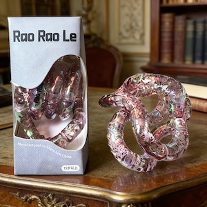 Op de afbeelding: Een doorzichtig, roze Rao Rao Le puzzelspeelgoed wordt naast de doos getoond. De doos heeft de tekst "Rao Rao Le" en "Manufactured in Shenzhen, China". De puzzel is een reeks in elkaar grijpende lussen.