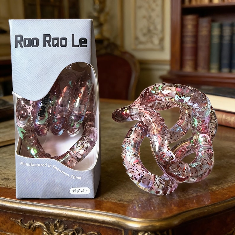 Op de afbeelding: Een doorzichtig, roze Rao Rao Le puzzelspeelgoed wordt naast de doos getoond. De doos heeft de tekst "Rao Rao Le" en "Manufactured in Shenzhen, China". De puzzel is een reeks in elkaar grijpende lussen.