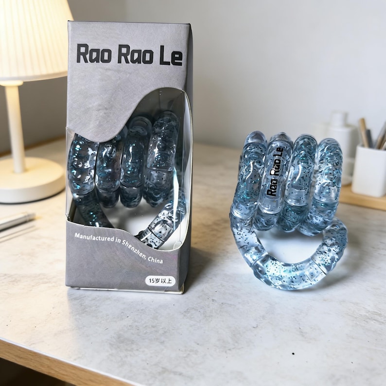 Op de afbeelding: Een helder, blauw, vuistvormig speelgoed met glitteraccenten, naast de originele verpakking. De verpakking leest "Rao Rao Le" en "Vervaardigd in Shenzhen, China". Het speelgoed is ontworpen om mee te manipuleren en te spelen.