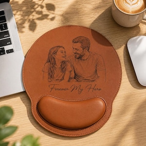 May include: A round, tan leather mouse pad with a built-in wrist rest. The surface features a dark brown line-art illustration of a smiling child and an adult looking at each other, with the text Forever My Hero printed below the image.