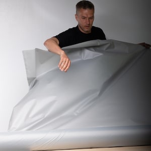 May include: A large roll of smooth, matte light gray material with a flexible, pliable texture. The material is unrolled to reveal a wide, continuous surface with subtle folds and creases across its expansive, uniform, and neutral-colored area.