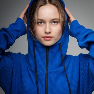 May include: A close-up of a blue hooded sweatshirt with a black zip and drawstrings. The sweatshirt has a textured pattern. The person is wearing the hood and holding it up with their hands.