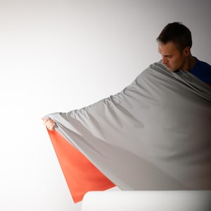 May include: A person holding a large, gray fabric with a bright orange underside. The fabric is draped and appears to be a sheet or blanket. The background is plain white.