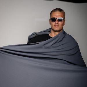 Op de afbeelding: Een man met een zonnebril en een zwart shirt, gedrapeerd met een grote, vloeiende, donkergrijze stof. De stof wordt uitgespreid, waardoor een dynamisch visueel effect ontstaat. De achtergrond is effen wit.