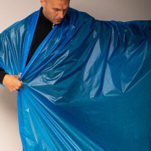 May include: A person holding up a large sheet of bright blue plastic material. The material is crinkled and appears to be rolled up at the bottom. The person is wearing a black turtleneck.