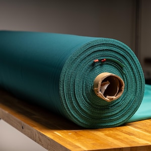 May include: A large roll of teal fabric sits on a wooden surface. The fabric is tightly wound, revealing the texture of the material. The core of the roll is a cardboard tube. A piece of the fabric is unrolled on the right.