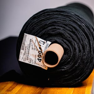 Op de afbeelding: Een grote rol zwarte stof ligt op een houten oppervlak. De stof is strak opgerold, waardoor een textuur zichtbaar is. Een label met tekst en een prijskaartje is bevestigd. Geschikt voor naaien of knutselen. De rol heeft een diameter van ongeveer 30 cm.