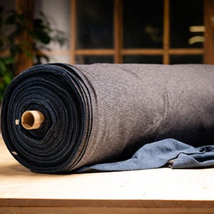 Peut inclure: Un grand rouleau de tissu, principalement dans les tons de gris et bleu marine, est posé sur une surface en bois. Le tissu est bien enroulé, révélant des couches de matière. Une partie du tissu est déroulée, montrant un tissu bleu plus doux.
