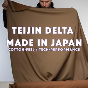 Pode incluir: Uma pessoa segurando um grande pedaço de tecido bege. O texto no tecido diz "TEIJIN DELTA MADE IN JAPAN COTTON-FEEL / TECH-PERFORMANCE". O tecido parece estar enrolado na parte inferior.