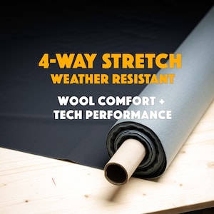 Op de afbeelding: Een rol donkergrijze stof met de tekst "4-WAY STRETCH WEATHER RESISTANT WOOL COMFORT + TECH PERFORMANCE" in geel en wit. De stof is uitgerold op een licht houten oppervlak.