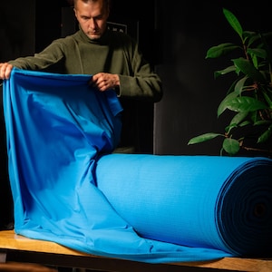 May include: A roll of bright blue fabric is displayed on a wooden surface. The fabric is being unfolded by a person wearing a green turtleneck sweater. The fabric appears to be a textured material.