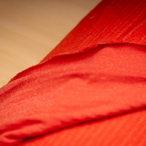 May include: Close-up of a vibrant red fabric, possibly a textile or material. The fabric is folded, revealing layers and textures. The colour is a bright, solid red, with subtle variations in tone due to the folds and lighting.
