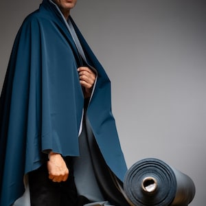 May include: A person draped in a teal-colored fabric, with a roll of matching fabric on a wooden surface. The fabric appears to be a smooth, possibly waterproof material. The background is a neutral gray.