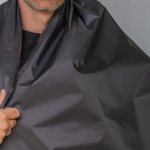 May include: A dark charcoal gray fabric featuring a visible grid-like ripstop pattern. The material has a smooth, slightly reflective surface texture and is draped in folds, showing a darker inner lining along the edge of the textile.